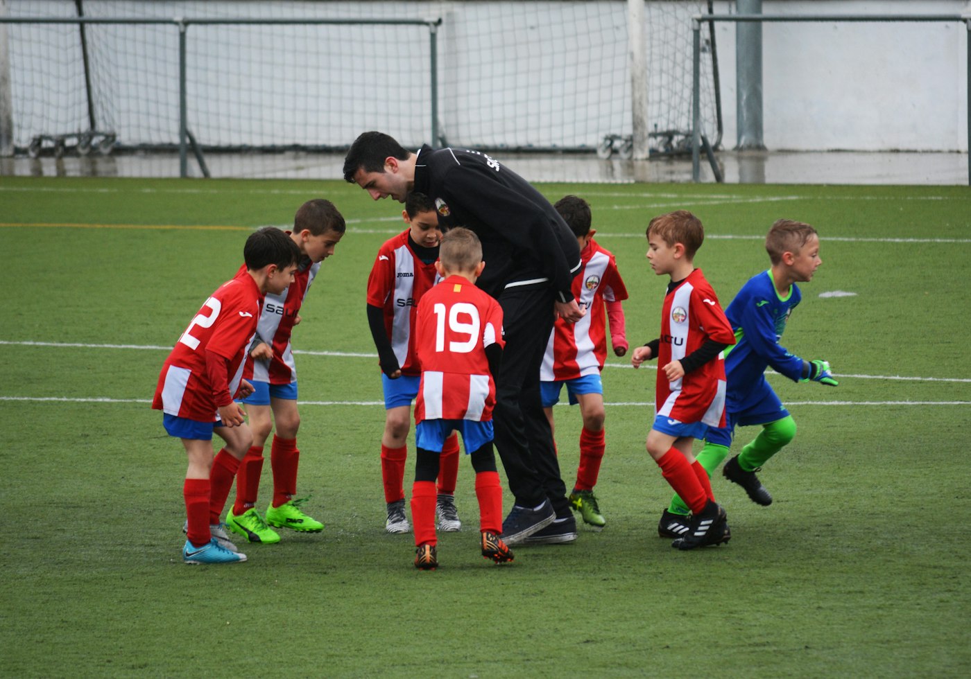 A youth soccer coach huddles with seven young players on a wet pitch