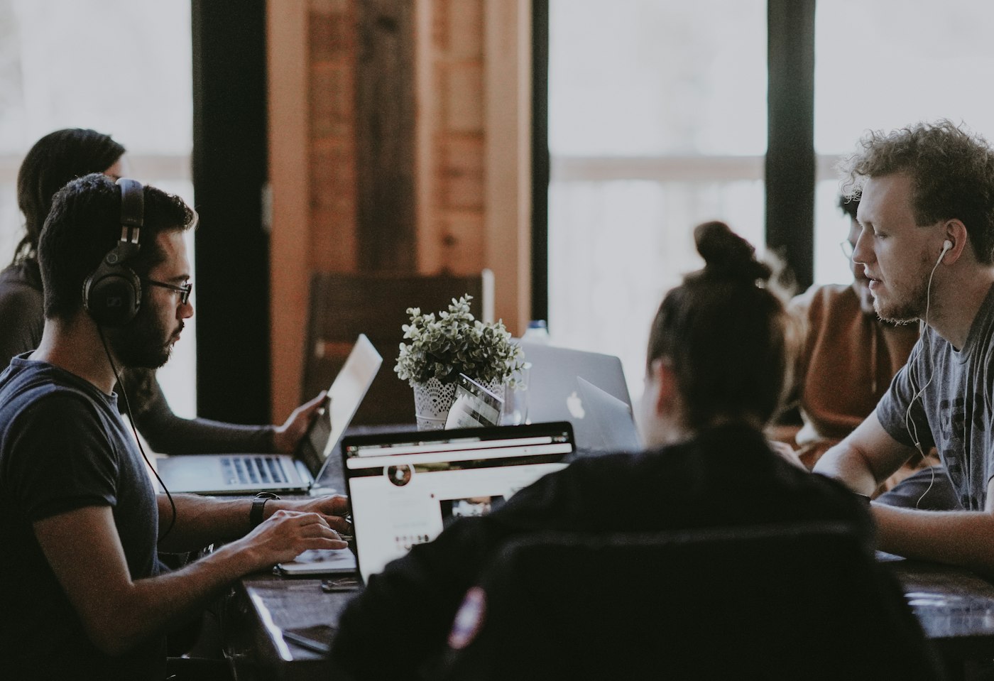 Five people working on laptops around a wooden table by a bright window